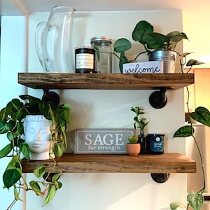 May include: Two rustic wooden shelves with black pipe brackets hold various decorative items. A clear pitcher, candles, and potted plants are displayed. One shelf has a sign that says "welcome", and the other says "SAGE for strength".