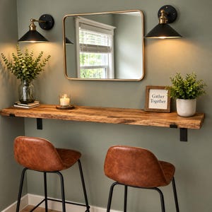 May include: A long, live-edge wooden floating shelf mounted with black metal brackets, featuring two brown faux leather bar stools, a rectangular mirror, two black wall sconces, a glass vase with greenery, a candle, and a sign reading Gather Together.