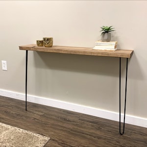 May include: A rustic wooden console table with black hairpin legs. The tabletop is made of light-colored wood and features a natural edge. Two gold decorative cups and a small potted plant with books are displayed on the table.