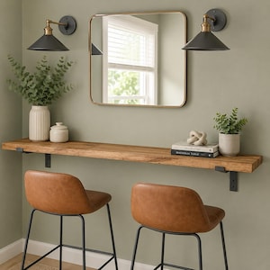 May include: A long wooden wall-mounted shelf with black metal brackets, two tan faux leather bar stools with black metal legs, a square mirror with a gold frame, two black wall sconces, two white vases with greenery, and two stacked books.