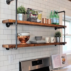 May include: Rustic, two-tiered kitchen shelving unit with dark wood shelves and black metal pipe supports. The shelves hold various kitchen items, including plants, copper mugs, plates, bowls, and bottles of water. The shelves are mounted on a white tiled wall.