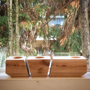 May include: Three wooden planter boxes, each with a square opening, are arranged in a row. The boxes are made of light brown wood and have a natural finish.