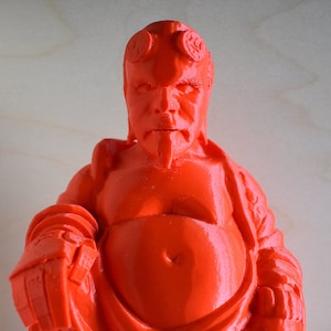 May include: A red 3D printed figurine of a smiling, bald man with horns, wearing a robe and sitting in a meditative pose. The figurine is a stylized representation of a popular comic book character.