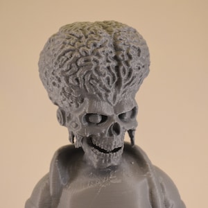 May include: A gray 3D printed figurine of a creature with a skull-like face and a brain-like head. The figurine is wearing a jacket.