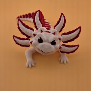 May include: A white and red axolotl figurine with a detailed design. The figurine has a stern expression and features red accents on its gills, back, and tail. The axolotl is posed on a solid orange background.