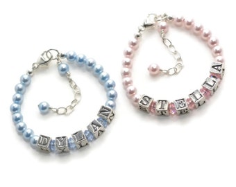 May include: Two beaded bracelets with silver-toned metal cube beads spelling the names DYLAN and STELLA. The DYLAN bracelet features light blue pearls and crystals, while the STELLA bracelet features light pink pearls and crystals.