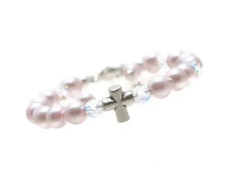 May include: A delicate ring featuring pale pink faux pearls and clear iridescent beads. A small silver-colored cross is centered on the ring. The ring is set against a white background.