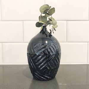 May include: A black ceramic vase with a textured, carved design. The vase has a narrow neck and a rounded body, and it holds a sprig of green foliage. The vase is sitting on a gray surface.
