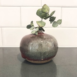 May include: A handcrafted, round ceramic vase with a speckled gray and red glaze. The vase holds a sprig of green eucalyptus leaves. The vase is displayed on a gray countertop against a white tiled background.