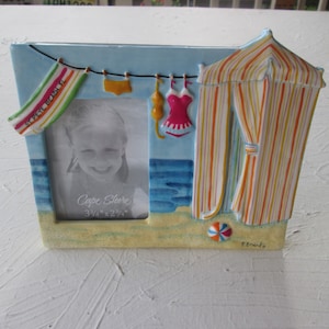 May include: A blue and yellow ceramic picture frame with a beach scene. The frame has a striped beach tent, a beach towel that says "Cape May Beach", and a beach ball. The frame is 3 1/2" x 2 1/4" and says "Cape Shore" on the bottom.