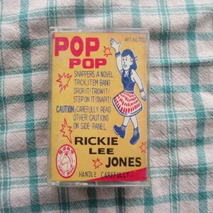 Op de afbeelding: Een gele en rode cassetteband met de tekst "Pop Pop" en "Rickie Lee Jones" op de voorkant. De band toont een cartoonmeisje in een rood-witte rok, dat een ster vasthoudt. De tekst "Snappers a Novel Trick.Item Bang Drop.It/Trowit! Step on It/Snapit!" staat op de band gedrukt. De tekst "Caution: Carefully read other cautions on side panel" staat ook op de band gedrukt. De band is gemarkeerd met "Horse Brand" en heeft de tekst "Handle Carefully."