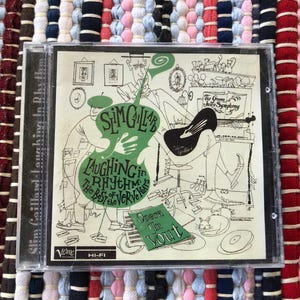 Peut inclure: Un boîtier de CD présentant l'album "Laughing in Rhythm" de Slim Gaillard. La pochette est une illustration en noir et blanc avec des accents verts, incluant le nom de l'artiste et le titre de l'album. Le boîtier de CD est posé sur un tapis tissé coloré.