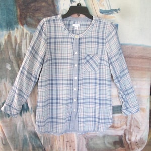 May include: A light blue plaid button-down shirt with long sleeves. The shirt features a rounded collar, a single chest pocket, and a button-up front. The plaid pattern includes white, red, and blue lines. The shirt is hanging on a black hanger.