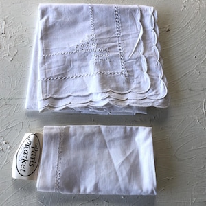 May include: Two white linen pillowcases, one folded with scalloped edges and embroidered details. The other pillowcase is folded with a simple stitched border. A label with the text "Paris Market" is also visible.