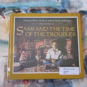 May include: A yellow book cover with the title "Sami and the Time of the Troubles" by Florence Parry Heide and Judith Heide Gilliland. The book cover features an illustration of a young boy sitting on a colorful rug.