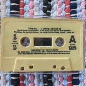 May include: A vintage cassette tape for the album "Apollo 18" by They Might Be Giants. The cassette is a light yellow color and is encased in a clear plastic case. The label features song titles and album information.