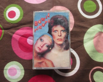May include: A cassette tape with a picture of David Bowie on the cover. The tape is titled "Pin Ups" and features a woman with a blue headband and a man with long red hair.
