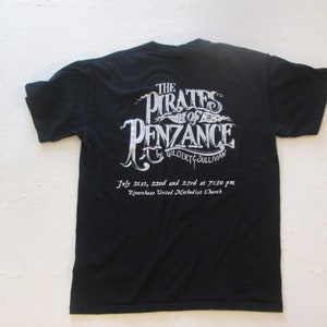 May include: Black t-shirt with white text advertising a performance of "The Pirates of Penzance" by Gilbert & Sullivan. The performance will take place on 21st, 22nd, and 23rd July at 7:30 pm at Riverchase United Methodist Church.