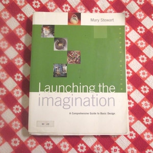 May include: A book titled "Launching the Imagination" with a green and white cover. The cover features the title in large, bold letters, along with the subtitle "A Comprehensive Guide to Basic Design." Small images and the author's name, Mary Stewart, are also visible.