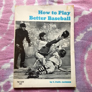 以下が含まれることがあります： 「How to Play Better Baseball」というタイトルのヴィンテージ本の表紙。野球の試合のシーンが描かれています。表紙には、キャッチャー、バッター、審判が描かれています。本はピンクと紫の模様の背景にあります。