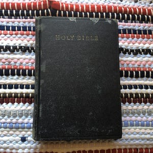 May include: A black hardcover Holy Bible with gold lettering, resting on a colorful woven rug. The book's cover shows signs of wear. The rug features stripes of red, white, blue, and other colors.