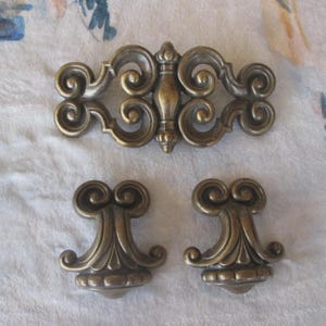 May include: Three antique bronze-toned furniture hardware pieces. The top piece is a long, ornate decorative element with scrollwork. Below are two matching smaller pieces, also with scroll designs, likely drawer pulls or knobs.