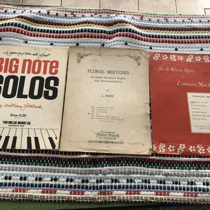 Può includere: Sono esposti tre libri di spartiti musicali vintage. I titoli includono "Big Note Solos", "Floral Sketches" e "To a Wild Rose". Il libro "Big Note Solos" presenta un'illustrazione di una tastiera di pianoforte. I libri sono su un tappeto a righe colorato.