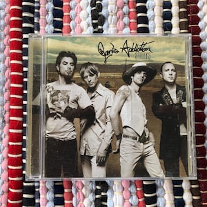 May include: A CD case featuring the band Jane's Addiction. The album cover shows four men in various outfits, with the band's name and album title, "Strays," displayed at the top. The background is a desert landscape.
