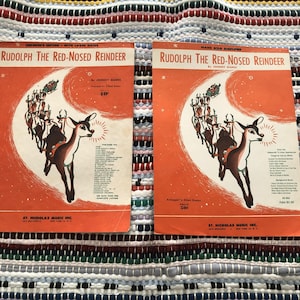 Puede incluir: Dos portadas de partituras antiguas de "Rudolph The Red-Nosed Reindeer" en naranja y blanco. Las portadas presentan una ilustración de Rudolph con una nariz brillante, un trineo y texto. La partitura es de St. Nicholas Music Inc.