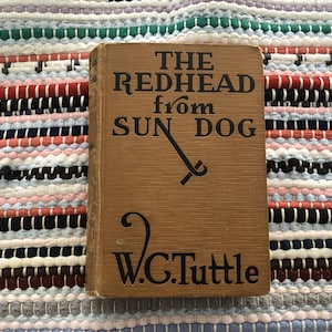 May include: A vintage book titled "THE REDHEAD from SUN DOG" by W.C. Tuttle. The book has a tan cover with black lettering and a stylized illustration. The book is resting on a colorful, striped textile.