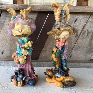 Peut inclure: Deux figurines décoratives de lapins de Pâques. L'un porte une robe rose et un chapeau lavande, tenant un bouquet de fleurs. L'autre porte une salopette et un chapeau, tenant des carottes. Les deux ont des détails et sont sur une surface en bois.
