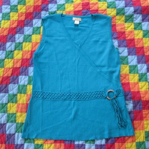 May include: A sleeveless turquoise top with a V-neck design. The top features a crocheted band at the waist with a wooden ring and fringe detail. The top is displayed on a colorful, patchwork quilt.