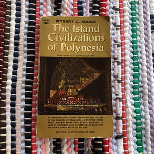 May include: A vintage paperback book titled "The Island Civilizations of Polynesia" by Robert C. Suggs. The cover is gold with black and brown illustrations. The book is set on a colorful, striped woven rug.