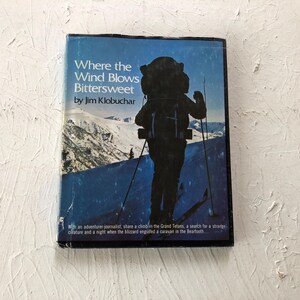 May include: A hardcover book titled Where the Wind Blows Bittersweet by Jim Klobuchar. The cover features a dark silhouette of a skier with a large backpack against a blue sky and snow-covered mountains. The text is printed in white font.