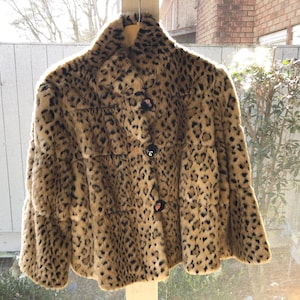 May include: A faux fur coat with a leopard print pattern. The coat has a round collar, three black buttons, and a flared hem. The coat is hanging on a wooden hanger.