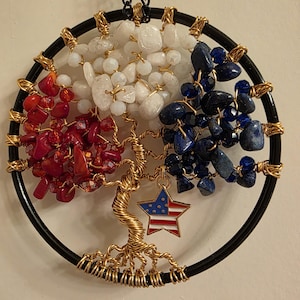 May include: A handcrafted wall hanging featuring a tree of life design. The circular frame is black, with the tree trunk and branches crafted from gold-colored wire. The tree's foliage is composed of red, white, and blue gemstones, with a star pendant featuring the American flag.