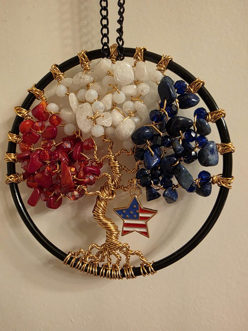 May include: A handcrafted wall hanging featuring a tree of life design. The circular frame is black, with the tree trunk and branches crafted from gold-colored wire. The tree's foliage is composed of red, white, and blue gemstones, with a star pendant featuring the American flag.
