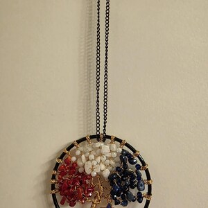 May include: A round, black metal dream catcher with red, white, and blue stones, and a gold wire tree design. A small star with an American flag design hangs from the tree. The dream catcher hangs from a black chain.
