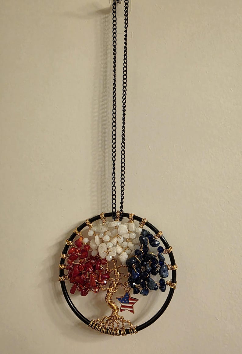 May include: A round, black metal dream catcher with red, white, and blue stones, and a gold wire tree design. A small star with an American flag design hangs from the tree. The dream catcher hangs from a black chain.