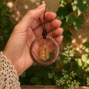 May include: A pendant necklace featuring a hammered copper ring and a central, wire-wrapped yellow citrine crystal point. The pendant is suspended from a dark brown leather cord, creating a contrast between the metallic texture and the stone.