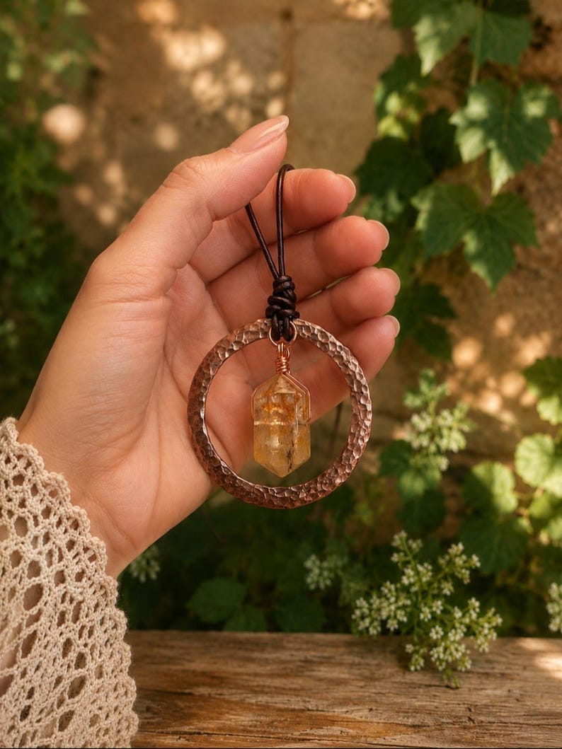 May include: A pendant necklace featuring a hammered copper ring and a central, wire-wrapped yellow citrine crystal point. The pendant is suspended from a dark brown leather cord, creating a contrast between the metallic texture and the stone.
