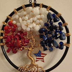 May include: A handcrafted tree of life wall hanging. The design features a black circular frame with a gold wire tree. The tree's branches are adorned with red, white, and blue stones, and a star charm with an American flag design.