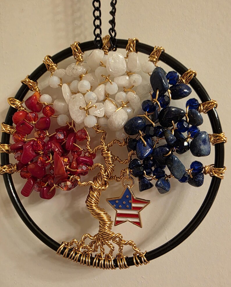 May include: A handcrafted tree of life wall hanging. The design features a black circular frame with a gold wire tree. The tree's branches are adorned with red, white, and blue stones, and a star charm with an American flag design.