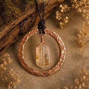 May include: A necklace featuring a hammered copper ring pendant suspended from a black leather cord. A clear, faceted crystal point is wrapped in copper wire and hangs from the center of the ring. The image includes the text #4.