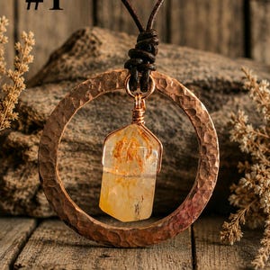 May include: A necklace featuring a hammered copper circular pendant with a central clear, yellow-tinted crystal point wrapped in copper wire. The pendant hangs from a dark brown leather cord. The image includes the text #1 in the top corner.