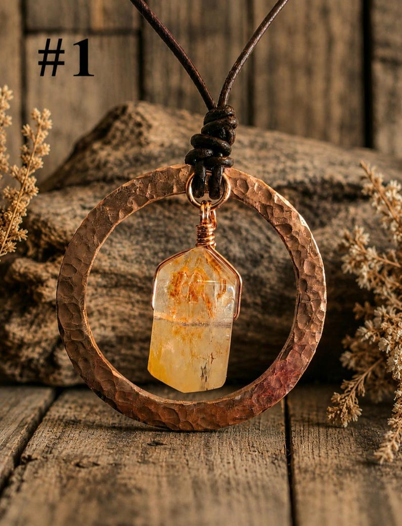 May include: A necklace featuring a hammered copper circular pendant with a central clear, yellow-tinted crystal point wrapped in copper wire. The pendant hangs from a dark brown leather cord. The image includes the text #1 in the top corner.