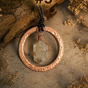 May include: A necklace featuring a hammered copper circular pendant suspended from a black cord. A clear, hexagonal crystal with internal golden inclusions is wire-wrapped in copper and hangs within the center of the circle. The text #5 is visible.