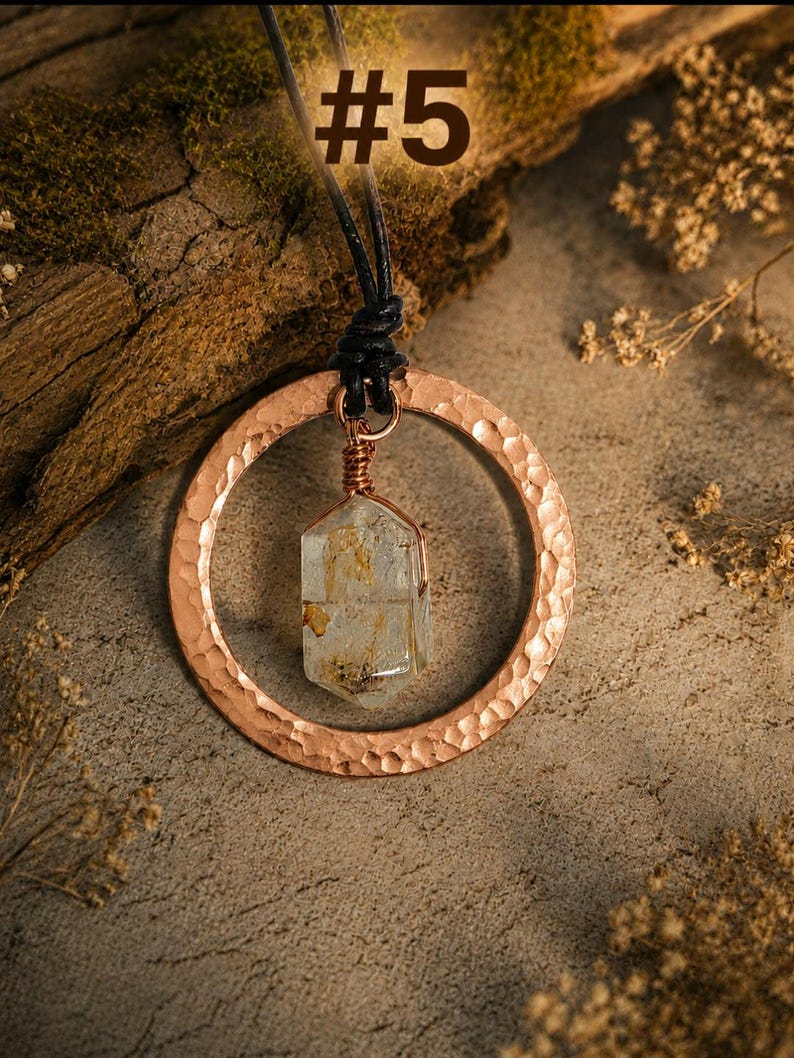 May include: A necklace featuring a hammered copper circular pendant suspended from a black cord. A clear, hexagonal crystal with internal golden inclusions is wire-wrapped in copper and hangs within the center of the circle. The text #5 is visible.