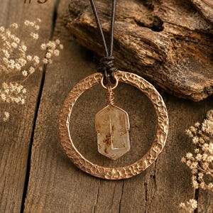 May include: A necklace featuring a hammered copper-colored circular pendant suspended from a dark brown leather cord. A clear, hexagonal-shaped crystal with brown inclusions is wire-wrapped and hangs inside the circle. The text #3 is visible.