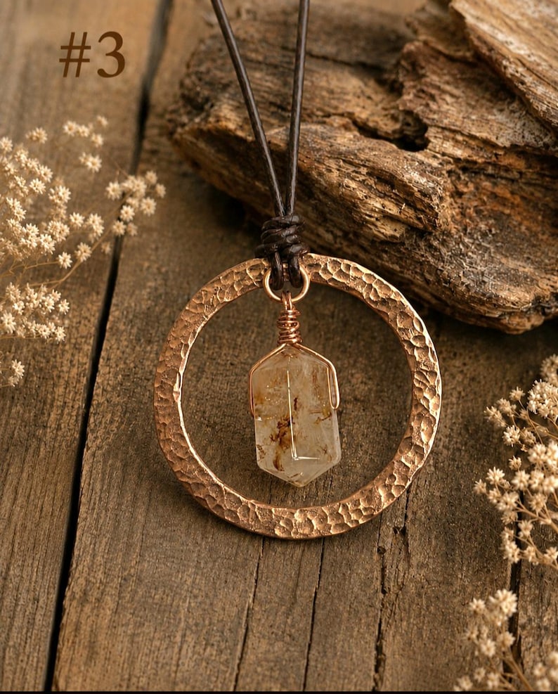 May include: A necklace featuring a hammered copper-colored circular pendant suspended from a dark brown leather cord. A clear, hexagonal-shaped crystal with brown inclusions is wire-wrapped and hangs inside the circle. The text #3 is visible.